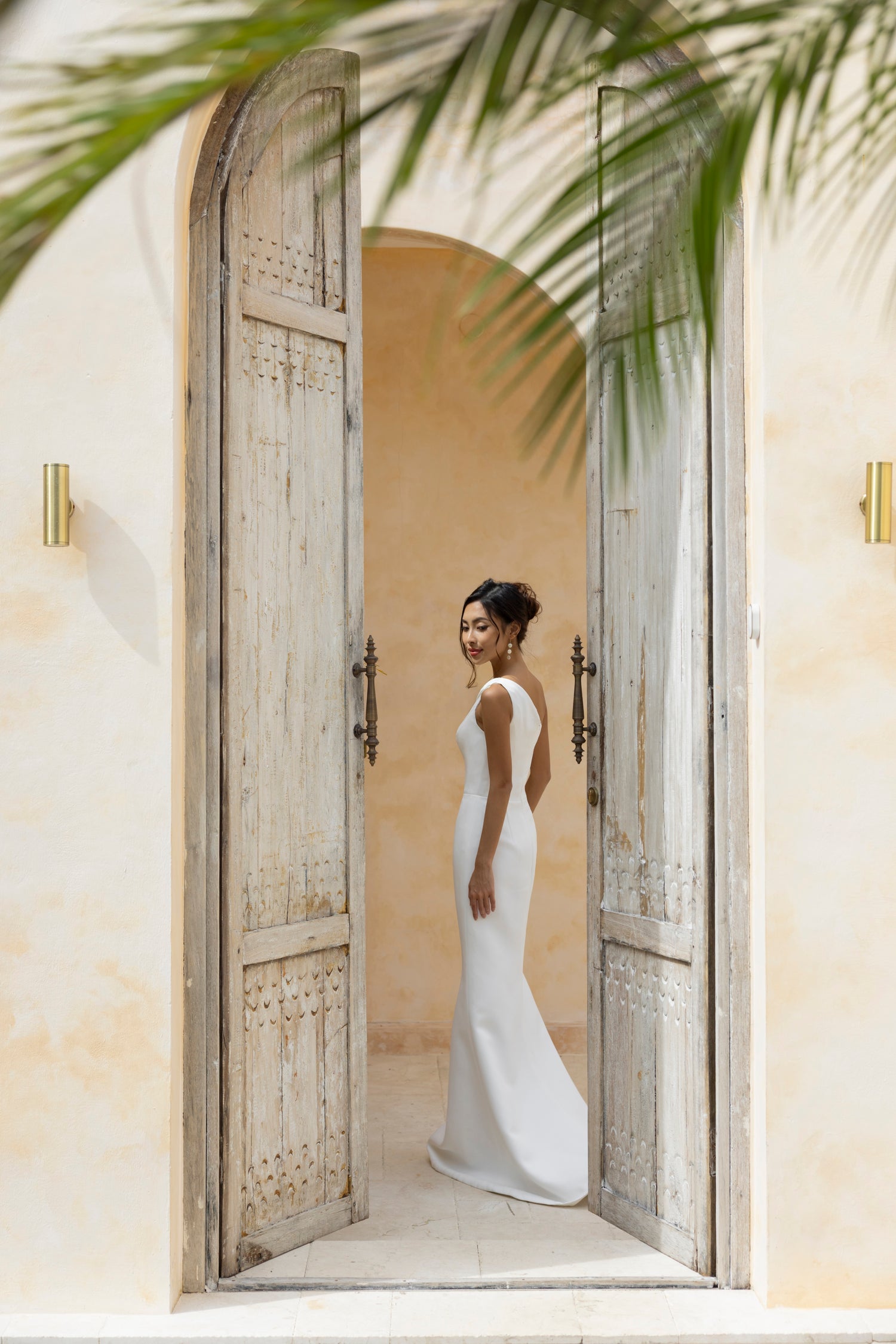 Bride wearing The Wolf and Lily Isla wedding dress in minimalist crepe with a deep V-back, standing in rustic archway